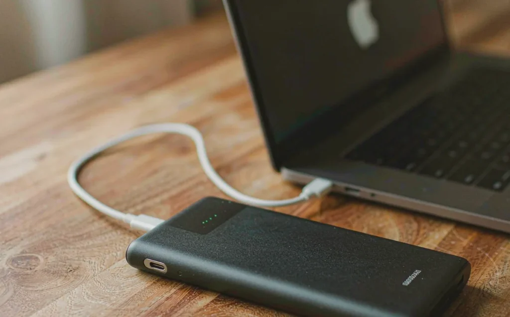 Black power bank with USB-C cable charging a laptop on a walnut desk